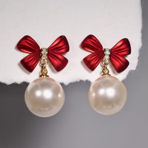 May include: A pair of red bow earrings with a pearl drop. The bows have a gold tone and are accented with clear rhinestones.