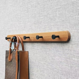 Op de afbeelding: Een houten wandrek met vijf zwarte metalen haken. Een bruine leren handtas hangt aan een van de haken.