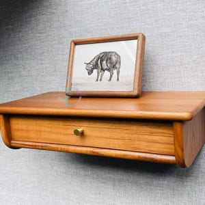 Op de afbeelding: Een houten wandplank met een lade en een ingelijste print van een zwart-witte koe. De plank is gemaakt van lichtbruin hout en heeft een eenvoudig, modern ontwerp.
