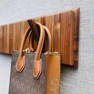 May include: Two brown leather handbags hanging on a wooden wall mounted rack. The rack is made of dark wood with a slatted design.