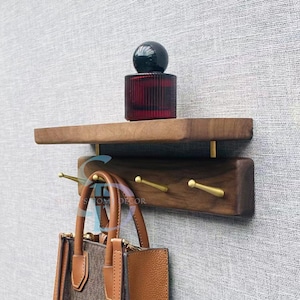 Könnte beinhalten: Ein hölzernes Wandregal mit dunklem Finish, mit einem Regal und einem unteren Abschnitt mit drei Messinghaken. Eine braune Handtasche hängt an den Haken. Eine rote Glasflasche und eine schwarze Kugel stehen auf dem Regal. Das Regal ist an einer strukturierten grauen Wand befestigt.