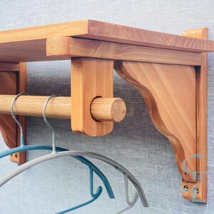 May include: A wooden wall-mounted shelf with a rod for hanging clothes. The shelf has a natural wood finish and is made of solid wood. The shelf is mounted on a wall with two brackets. The shelf is approximately 24 inches wide and 12 inches deep.