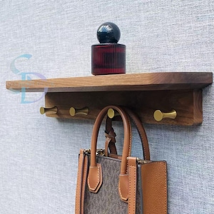 Peut inclure: Une étagère en bois avec trois crochets en laiton, tenant un sac à main marron. Une bouteille décorative rouge et noire est posée sur l'étagère. L'étagère est fixée à un mur gris texturé.