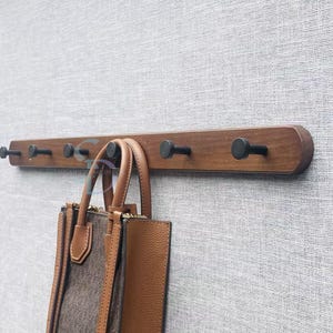 Könnte beinhalten: Eine horizontale Wandgarderobe aus Holz mit sechs gleichmäßig verteilten, mattschwarzen zylindrischen Haken. Eine braune Ledertasche mit strukturierter Oberfläche und zwei Tragegriffen hängt an einem der Haken der dunklen Holzleiste.