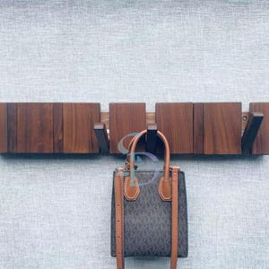 May include: A wooden wall-mounted coat rack with a dark brown finish. The rack has several folding hooks and is holding a brown and gray handbag with tan leather accents. The background is a light gray textured wall.
