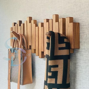 Puede incluir: Perchero de pared de madera con un diseño único. El perchero presenta listones de madera verticales de diferentes alturas. Un bolso de mano de cuero marrón y una bufanda con un patrón negro y marrón cuelgan de los ganchos. La pared es de color gris claro.