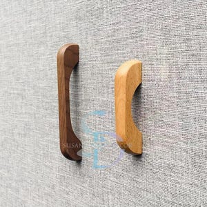 Könnte beinhalten: Zwei Holzschrankgriffe. Einer ist dunkelbraun, der andere hellbraun. Beide sind gebogen und haben einen Ausschnitt zum Greifen. Die Griffe sind auf einer grau strukturierten Oberfläche montiert.