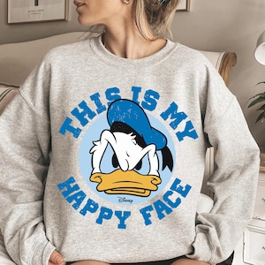 May include: A light gray sweatshirt featuring a graphic of Donald Duck with the text "THIS IS MY HAPPY FACE" in blue. The Disney logo is at the bottom of the graphic. The sweatshirt has long sleeves.