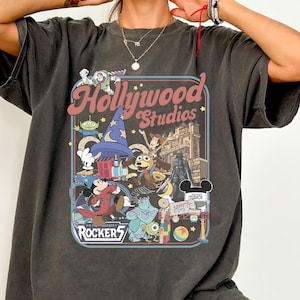 Peut inclure: Un t-shirt gris anthracite à manches courtes avec une impression graphique colorée portant le texte Hollywood Studios et Rockers. Le motif comprend divers personnages animés, un chapeau de magicien et l'architecture du parc dans un cadre rectangulaire.