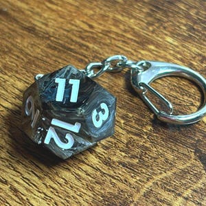 May include: A twenty-sided die keyring featuring a marbled black and white resin finish with bold white numbers. The die is attached to a silver-toned metal chain and a circular carabiner clip, creating a functional accessory for gaming enthusiasts.