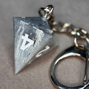 Op de afbeelding: Een vierzijdige, piramidevormige dobbelsteen-sleutelhanger met een gemarmerde grijs-witte harsafwerking en witte cijfers. De dobbelsteen is bevestigd aan een zilverkleurige metalen ketting en een ronde ring, met het cijfer vier prominent aan de voorzijde.