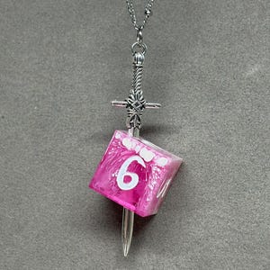 Puede incluir: Un collar de metal plateado con un colgante en forma de espada que atraviesa un dado de seis caras translúcido, jaspeado en rosa y blanco. El dado muestra el número seis en una fuente blanca grande en su cara frontal, suspendido de una cadena.
