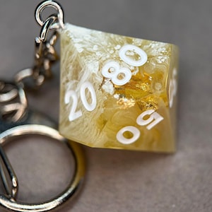 Könnte beinhalten: Ein Schlüsselanhänger mit einem polyedrischen Würfel aus durchscheinendem gelbem Harz, der weiße Zifferngravuren sowie weiße blumenartige Einschlüsse und Blattgoldakzente aufweist. Der Würfel ist an einer silberfarbenen Metallkette befestigt.