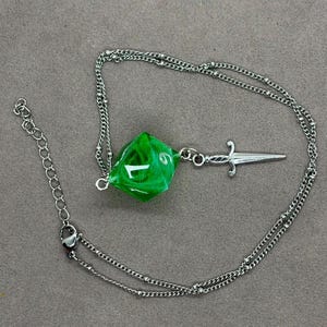 Puede incluir: Un collar de cadena de metal plateado con un colgante de dado de veinte caras verde, translúcido y facetado. El dado está unido a un pequeño dije de espada plateado, y la cadena incluye un cierre de mosquetón y una cadena de extensión.