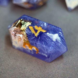 Puede incluir: Un dado poliédrico de múltiples caras hecho de resina púrpura translúcida con nubes blancas arremolinadas y copos de pan de oro incrustados. El dado presenta un número cuatro de color dorado grabado en su cara superior en fuente serif.