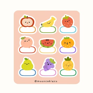 May include: A sheet of fruit stickers featuring an apple, banana, strawberry, peach, watermelon, orange, pear, grapes, and pineapple. Each sticker has a smiling face and a blank label. The background is a pale peach colour.