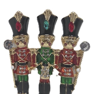 May include: A gold-toned brooch featuring three toy soldier figures. Each soldier wears a black hat with a jewel, a red or green jacket, and black boots. The brooch is embellished with rhinestones and has a festive, holiday theme.