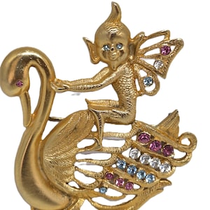 Peut inclure: Une broche en métal doré représentant une petite créature ailée assise sur un cygne. La créature a des yeux bleus et le cygne a un œil rose. Les ailes et les plumes de la queue du cygne sont ornées de petits strass ronds multicolores.