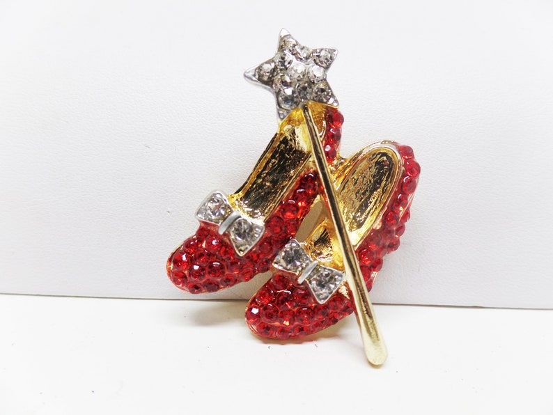 Puede incluir: Un broche de zapato rojo rub&iacute; con una varita dorada y una estrella en la parte superior. El broche est&aacute; hecho de diamantes de imitaci&oacute;n y es un objeto de colecci&oacute;n popular.