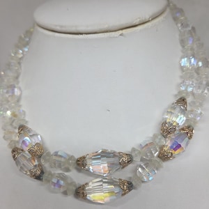 以下が含まれることがあります： イリデセントアクセントが付いた、2連のクリアクリスタルネックレス。 ネックレスは、ファセットビーズと、金色の金属アクセントが付いた、より大きな楕円形のクリスタルが特徴です。 ネックレスは白いジュエリーバストに展示されています。