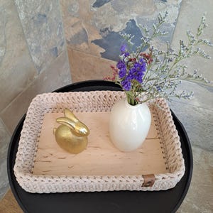 Könnte beinhalten: Ein beigefarbenes gehäkeltes Tablett mit einer Holzbasis, verziert mit einer goldenen Hasenfigur und einer weißen Vase mit violetten Blumen. Das Tablett steht auf einem schwarzen Tisch.