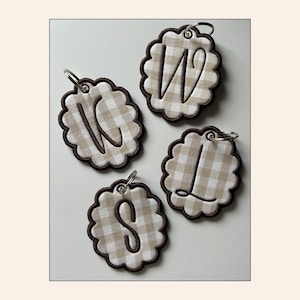 May include: Four scalloped-edge circular tags featuring a beige and white gingham pattern. Each tag is embroidered with a single dark brown letter—W, K, S, or L—in a cursive script. Each tag includes a silver-toned metal split ring for attachment.