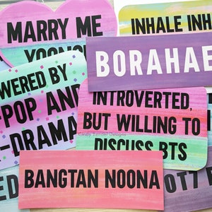 May include: A collection of colorful signs with various phrases in black text. The signs have a painted look with pastel backgrounds. Phrases include "Marry Me", "Powered by K-Pop and Drama", and "Introverted, but willing to discuss BTS".