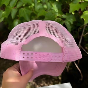 May include: A light pink trucker hat with a mesh back and adjustable snap closure. The hat's brim and front panel are solid pink, while the back is a breathable mesh. The adjustable strap allows for a custom fit.