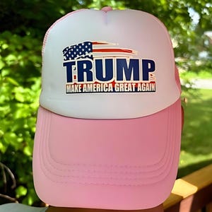 May include: A pink and white trucker hat with the word "TRUMP" in blue block letters, above the phrase "MAKE AMERICA GREAT AGAIN". The hat features a graphic of the American flag.