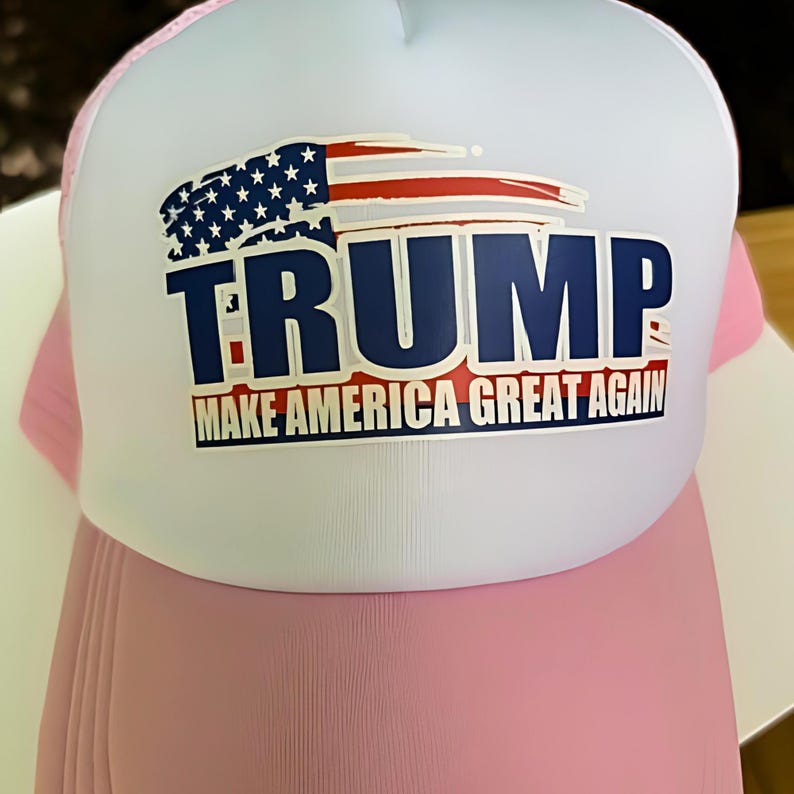 May include: A trucker hat with a white front panel and a pink mesh back. The hat features the word "TRUMP" in navy blue block letters, with an American flag graphic above it. The text "MAKE AMERICA GREAT AGAIN" is below the name.
