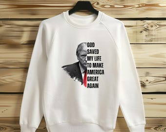 May include: White sweatshirt with a black and white image of a man with the text "God Saved My Life To Make America Great Again".