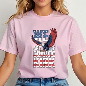 Puede incluir: Camiseta rosa claro con un &aacute;guila calva americana con las alas extendidas. El texto "WE ARE CHARLIE KIRK" se muestra en rojo, blanco y azul, reflejando la bandera estadounidense. La camiseta es de estilo cuello redondo cl&aacute;sico.