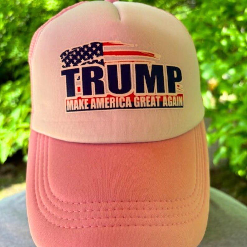 May include: Pink and white trucker hat with the word "TRUMP" in navy blue block letters and an American flag graphic. The text "MAKE AMERICA GREAT AGAIN" is below the word "TRUMP". The hat has a mesh back.