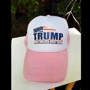 May include: Pink and white trucker hat with the word "TRUMP" in blue block letters and the phrase "MAKE AMERICA GREAT AGAIN" in red. The hat features an American flag design.