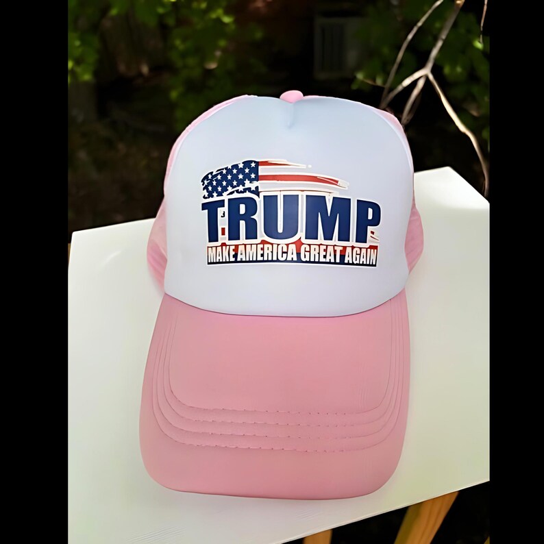 May include: Pink and white trucker hat with the word "TRUMP" in blue block letters and the phrase "MAKE AMERICA GREAT AGAIN" in red. The hat features an American flag design.