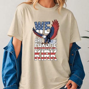 Puede incluir: Camiseta beige con el texto "WE ARE CHARLIE KIRK" en un dise&ntilde;o patri&oacute;tico con un &aacute;guila con las alas extendidas. El texto incorpora los colores de la bandera americana: rojo, blanco y azul.