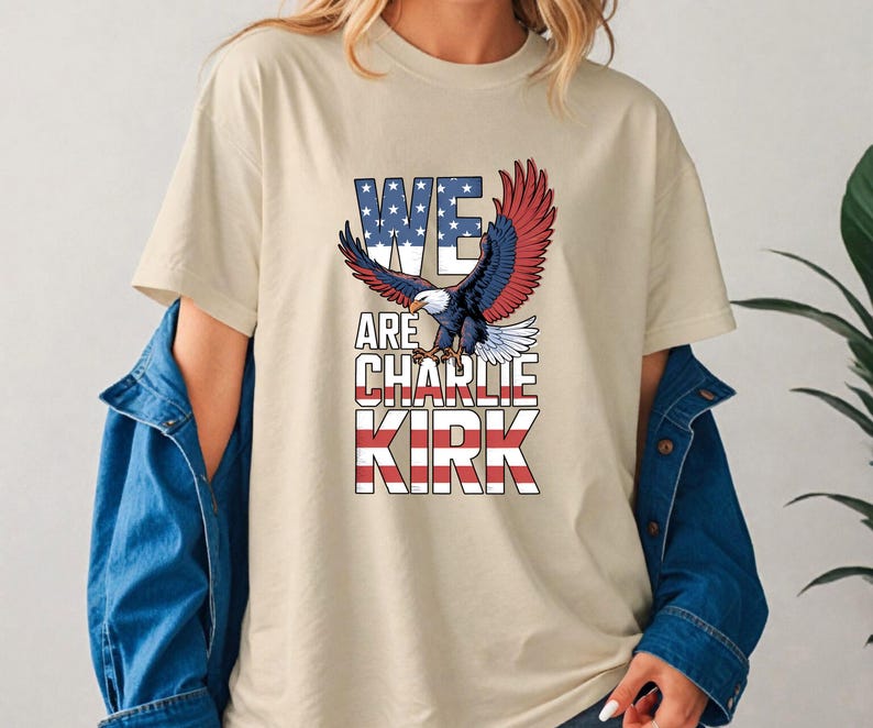 Puede incluir: Camiseta beige con el texto "WE ARE CHARLIE KIRK" en un dise&ntilde;o patri&oacute;tico con un &aacute;guila con las alas extendidas. El texto incorpora los colores de la bandera americana: rojo, blanco y azul.