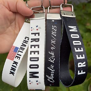 Puede incluir: Cuatro llaveros con herrajes plateados. Uno presenta la bandera estadounidense y el nombre "Charlie Kirk". Otro dice "FREEDOM" con estrellas. Un tercero tiene el nombre "Charlie Kirk 9/10/2025". El último dice "FREEDOM" en blanco sobre negro.