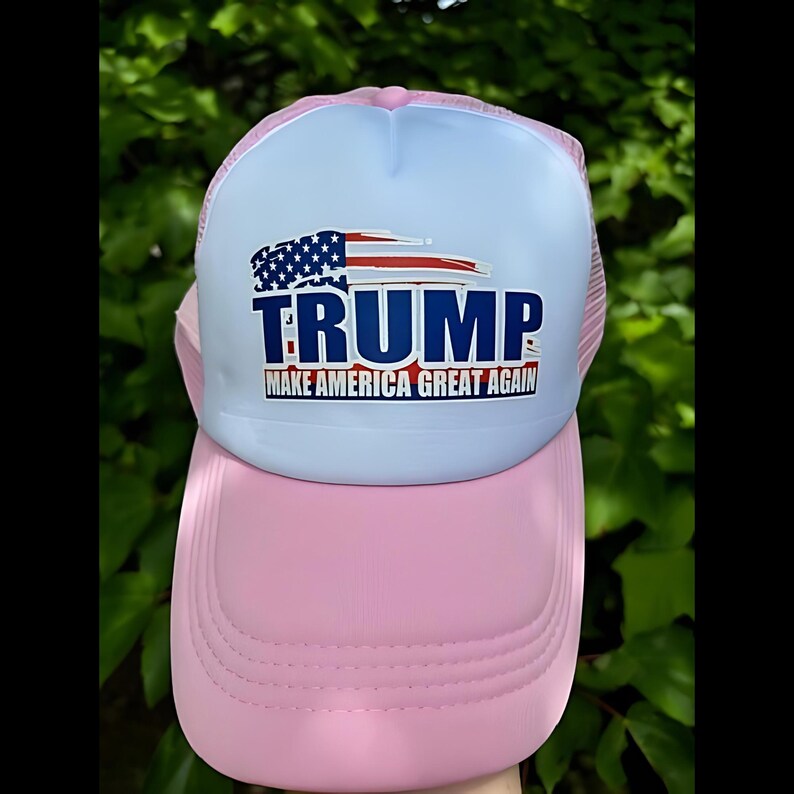 May include: A pink and light blue trucker hat with the word "TRUMP" in navy blue block letters. The hat features an American flag design and the text "MAKE AMERICA GREAT AGAIN" in red and white.