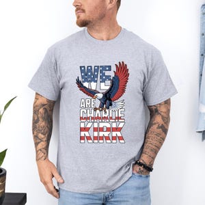Puede incluir: Camiseta gris claro con un &aacute;guila de alas rojas, blancas y azules y el texto "WE ARE CHARLIE KIRK". El &aacute;guila est&aacute; sobre el texto, que est&aacute; dise&ntilde;ado para parecerse a la bandera estadounidense.
