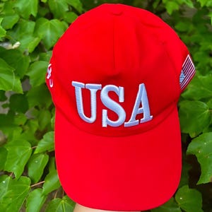 May include: A bright red baseball cap with the word "USA" embroidered in white across the front. The cap features a curved brim and a small American flag patch on the side. The background is a lush green foliage.