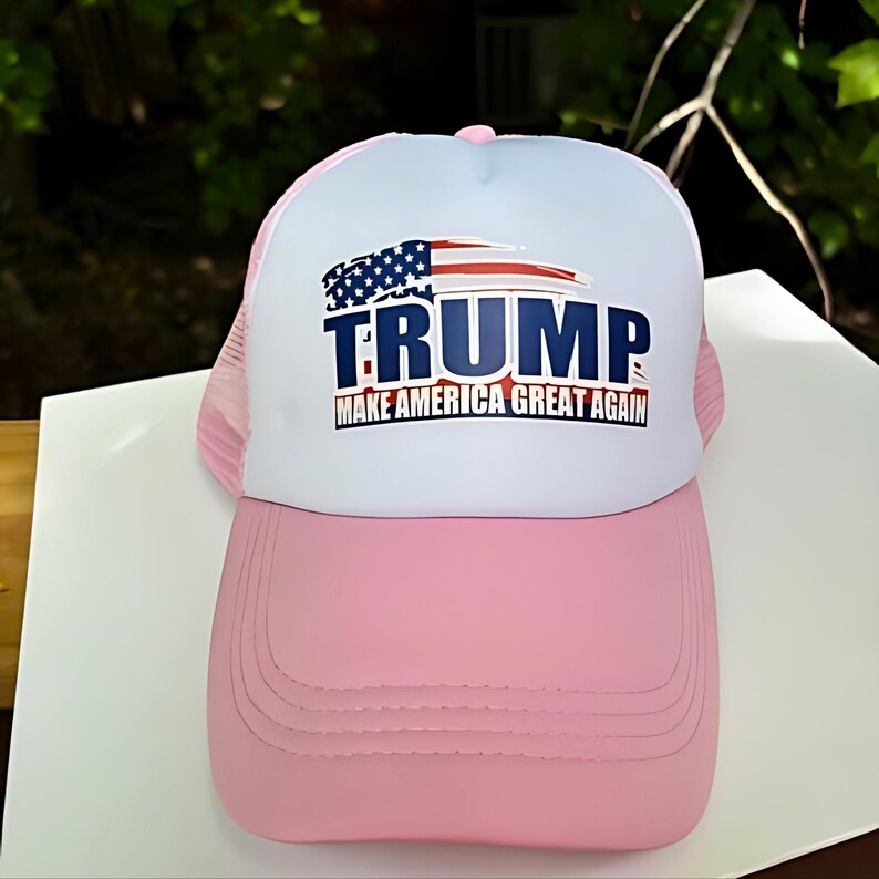 May include: Pink and white trucker hat with the text "TRUMP" and the phrase "MAKE AMERICA GREAT AGAIN" printed on the front. The hat features a mesh back and a curved brim.