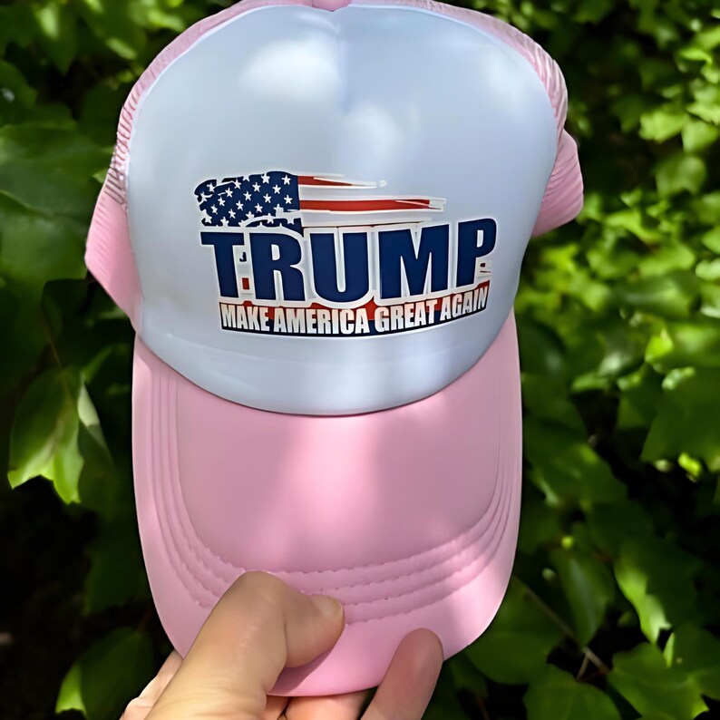 May include: Pink and white trucker hat with the word "TRUMP" in blue block letters. The hat features an American flag graphic and the phrase "MAKE AMERICA GREAT AGAIN" in white text.