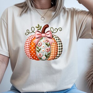 以下が含まれることがあります： ピンクのリボンが付いたカボチャのグラフィックが中央に描かれたベージュのTシャツ。カボチャは、オレンジ色の水玉模様、オレンジ色のギンガムチェック、緑色のギンガムチェック、そして花柄の刺繍が施された中央のパネルなど、さまざまなパターンのセグメントで構成されています。
