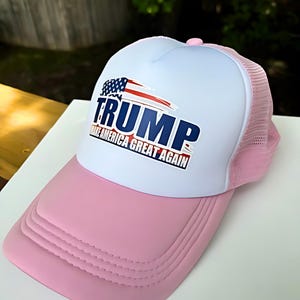 May include: Pink and white trucker hat with the word "TRUMP" in blue and the phrase "MAKE AMERICA GREAT AGAIN" in red. The hat features a white front panel, a pink brim, and a pink mesh back.