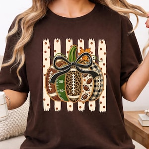 Könnte beinhalten: Ein dunkelbraunes Kurzarm-T-Shirt mit einer zentralen Grafik eines Kürbisses, der aus verschiedenen Mustern wie Football-Texturen, Leopardenmuster und schwarz-weißem Karomuster besteht, gekrönt von einer schwarzen Schleife.