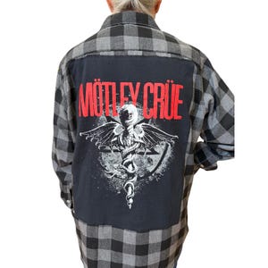 Könnte beinhalten: Ein grau-schwarz kariertes Flanellhemd mit einem schwarzen Einsatz auf dem Rücken. Auf dem Einsatz ist der Bandname "Mötley Crüe" in Rot abgebildet, darunter eine weiße Grafik mit einem Totenkopf, Flügeln und einer Schlange.