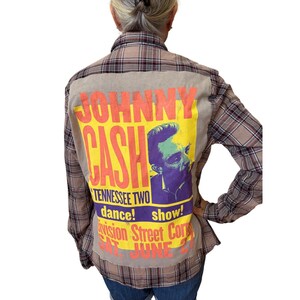 May include: A brown plaid flannel shirt with a large graphic on the back featuring Johnny Cash. The graphic is on a tan background with yellow and purple text and a portrait of Johnny Cash.