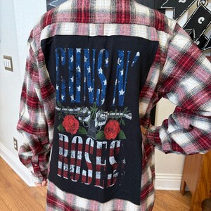 Puede incluir: Una camisa de franela a cuadros rojos, blancos y negros con un panel negro en la espalda. El panel presenta el texto "GUNS N' ROSES" con un diseño de pistolas y rosas.
