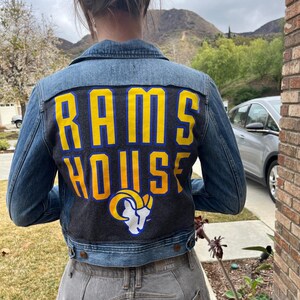 Puede incluir: Una chaqueta vaquera azul con un gráfico Rams House negro y amarillo en la espalda. El gráfico presenta un logotipo de cabeza de carnero.